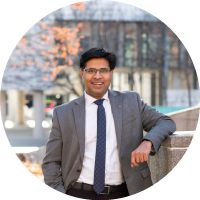 Headshot of Aravind Chandrasekaran wearing glasses and a suit with a tie, smiling outdoors.