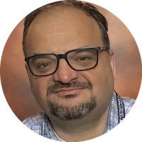 Headshot of Ibrahim Haskara wearing glasses and a plaid shirt, smiling gently against a studio backdrop.