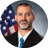 Headshot of Dr. Joseph B. Lyons wearing glasses and a dress shirt, smiling against a studio background.