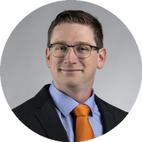 Headshot of Mark Weir wearing glasses and a suit with an orange tie, smiling against a neutral gray background.