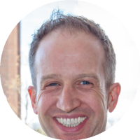 Headshot of Michael Rayo wearing a suit jacket and tie, smiling in front of a blurred outdoor/architectural background.