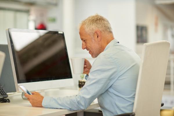 A man in an office sitting at a desk looking at his phone and drinking coffee
