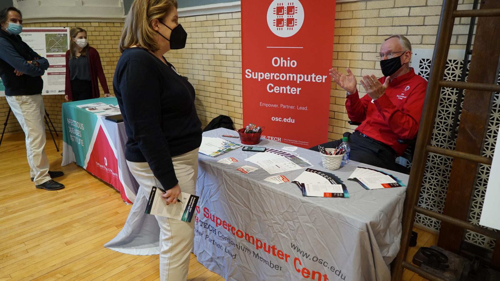 A woman speaks with a man who is seated at an Ohio Supercomputing Center table at the TDAI Fall Forum