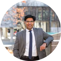 Headshot of Aravind Chandrasekaran wearing glasses and a suit with a tie, smiling outdoors.