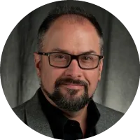 Headshot of Duane Detwiler wearing glasses and a blazer with a trimmed beard, against a studio background.