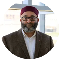 Headshot of Ayaz Hyder wearing glasses and a jacket, standing indoors.
