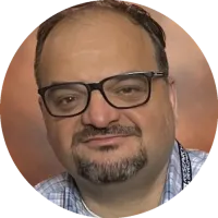 Headshot of Ibrahim Haskara wearing glasses and a plaid shirt, smiling gently against a studio backdrop.