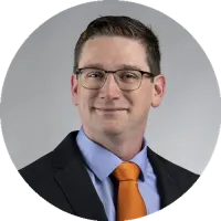 Headshot of Mark Weir wearing glasses and a suit with an orange tie, smiling against a neutral gray background.